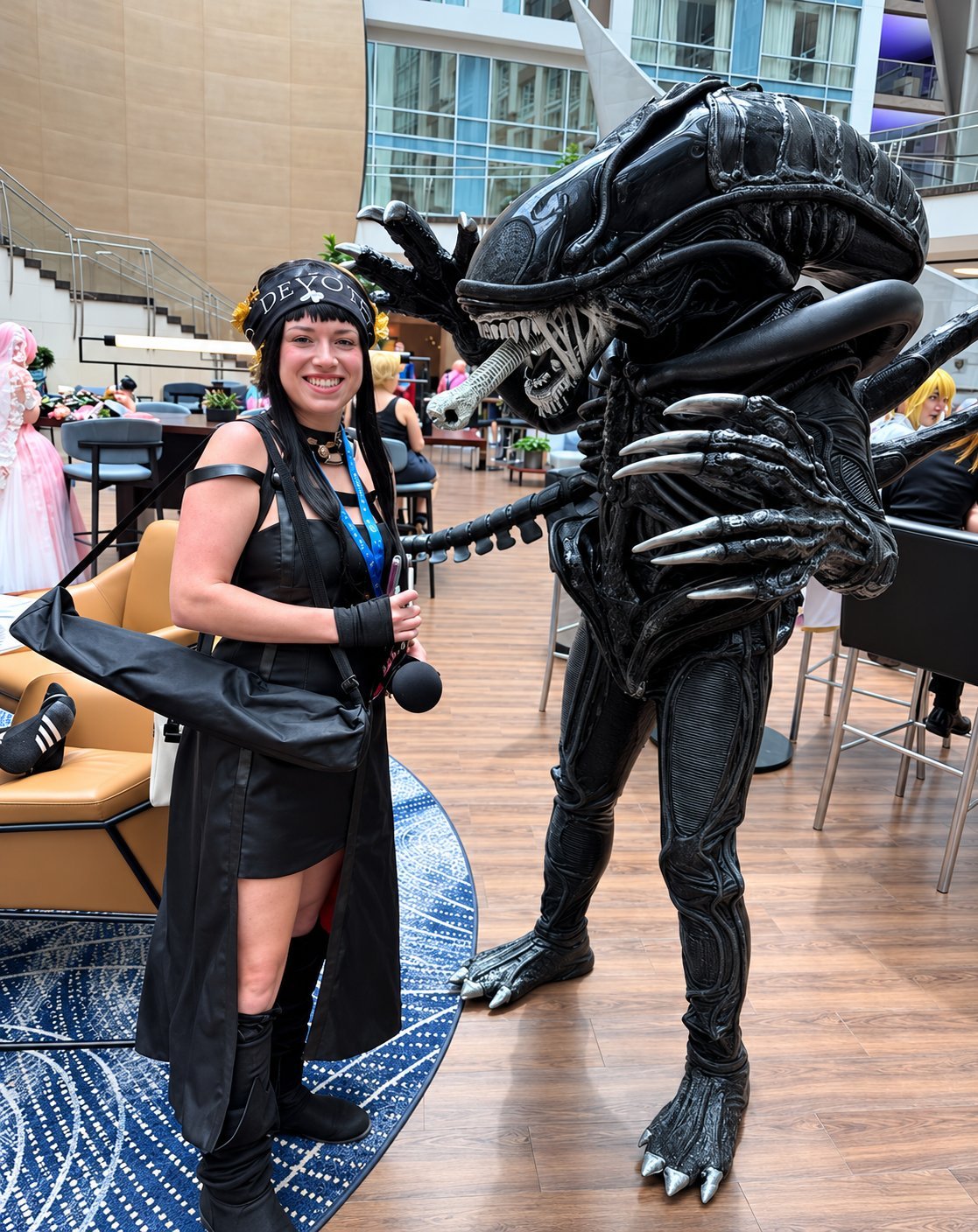 Woman in black alien costume poses with life-size xenomorph figure at indoor convention or event