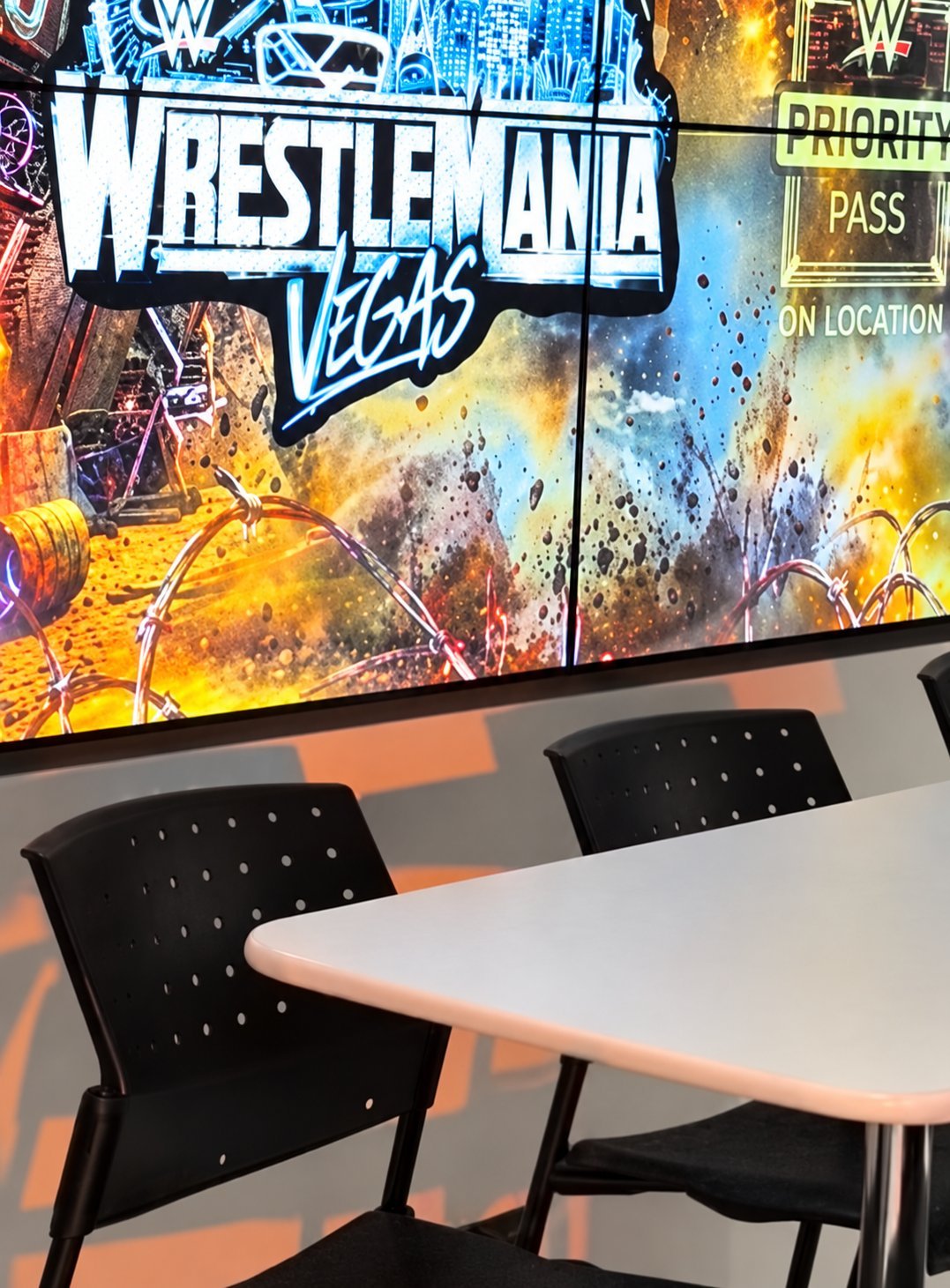 WrestleMania Vegas signage displayed on a large screen above a table with black chairs in a WWE event venue