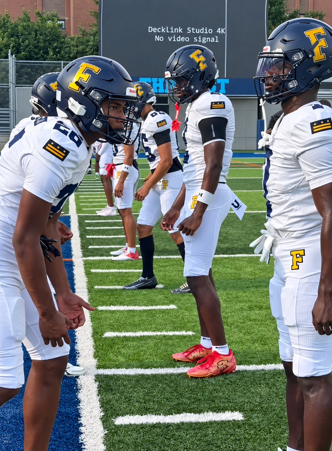 Football players in white jerseys and navy helmets with gold F logos huddle together on a football field during practice