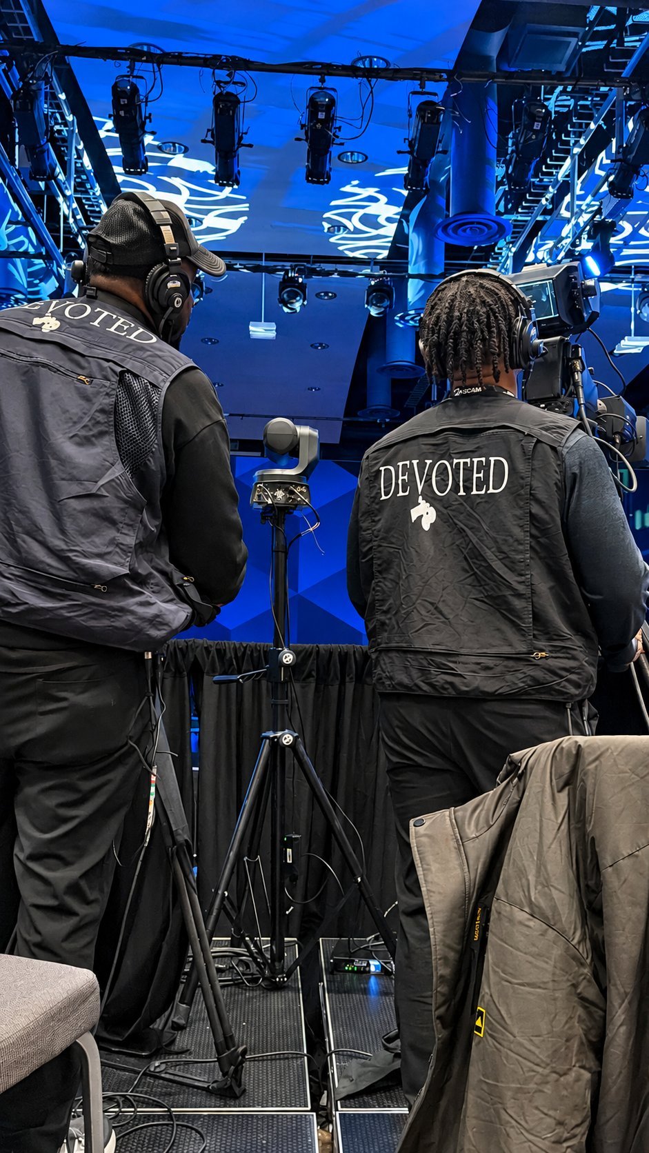 Two crew members wearing DEVOTED jackets operate camera equipment on a professional film set with blue lighting and overhead rigs